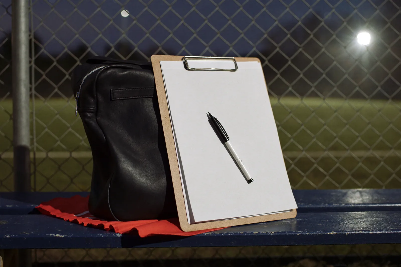 A blank clipboard and pen beside a kitbag on a pitchside bench under floodlights