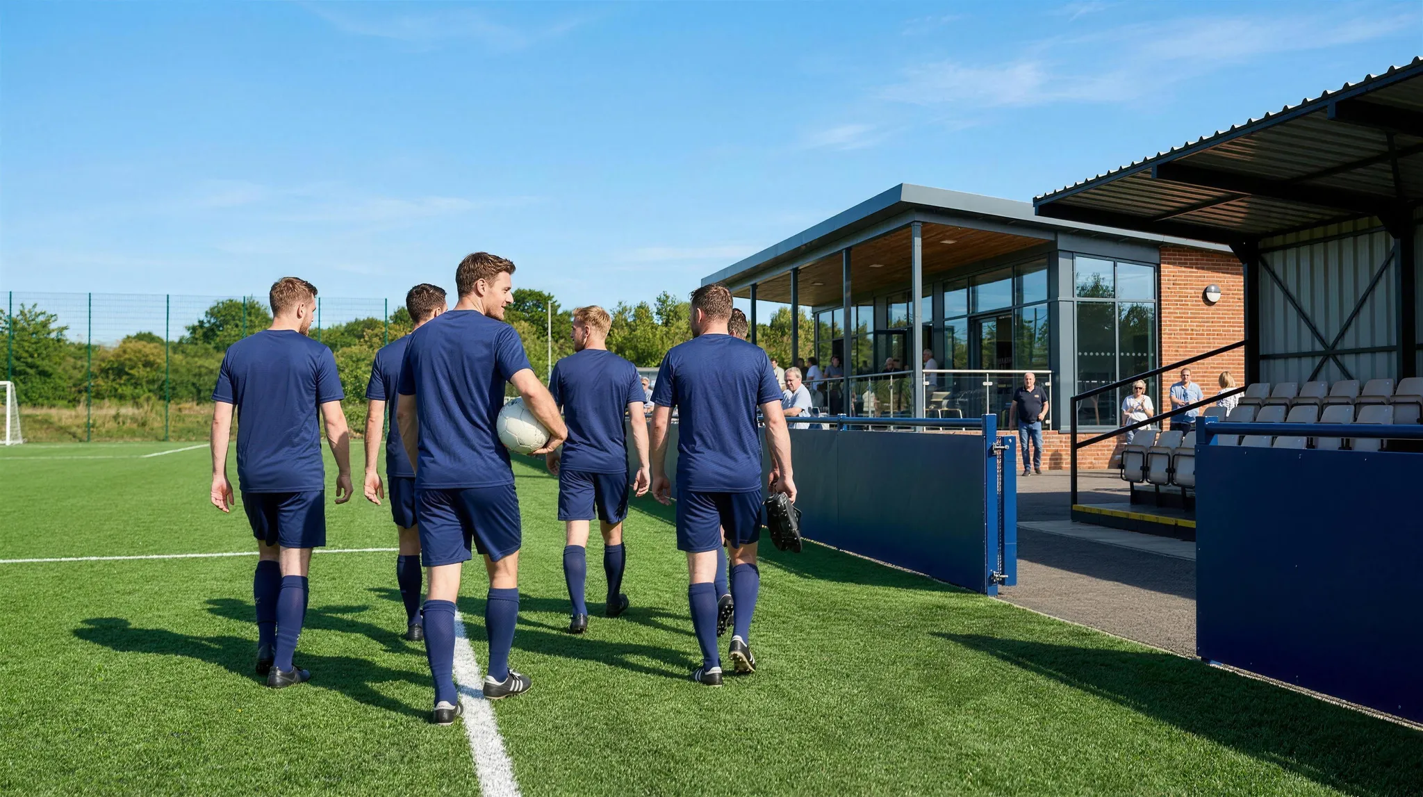Players in navy kit walking off a bright 3G pitch toward a modern clubhouse with people on the terrace