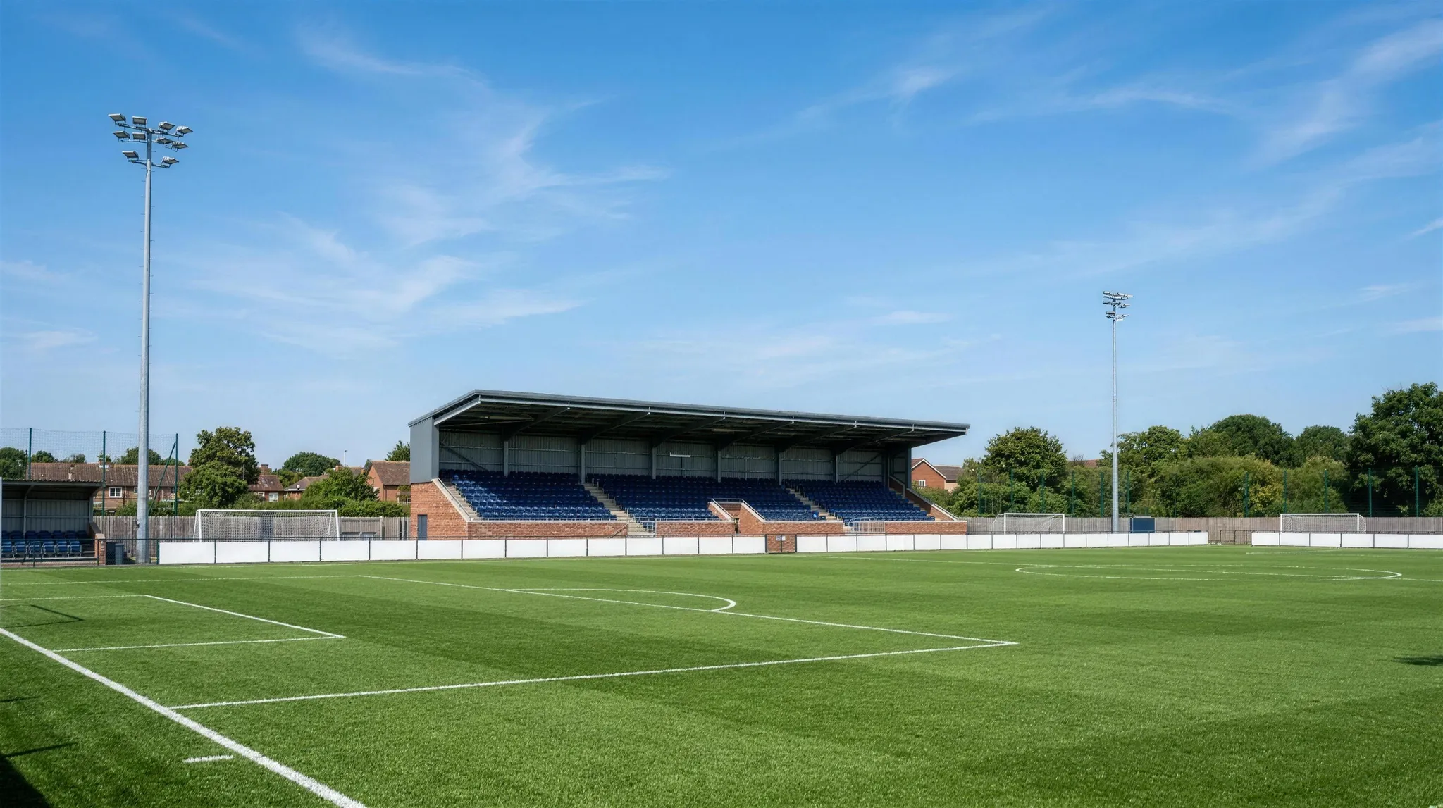 An empty non-league football pitch with a covered seated stand under a clear blue sky