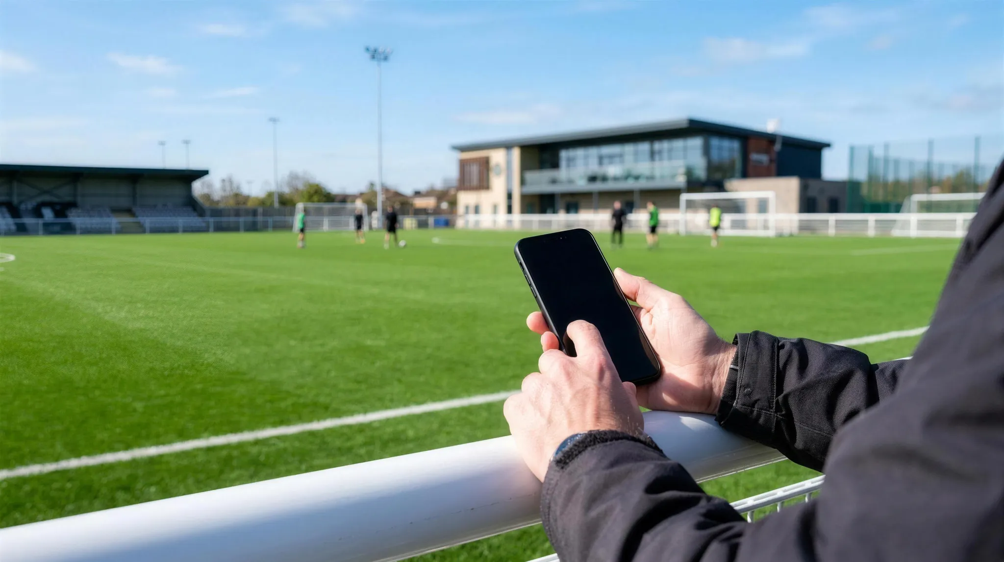Leaning on the rail with a phone in hand, a training session under way on a bright 3G pitch