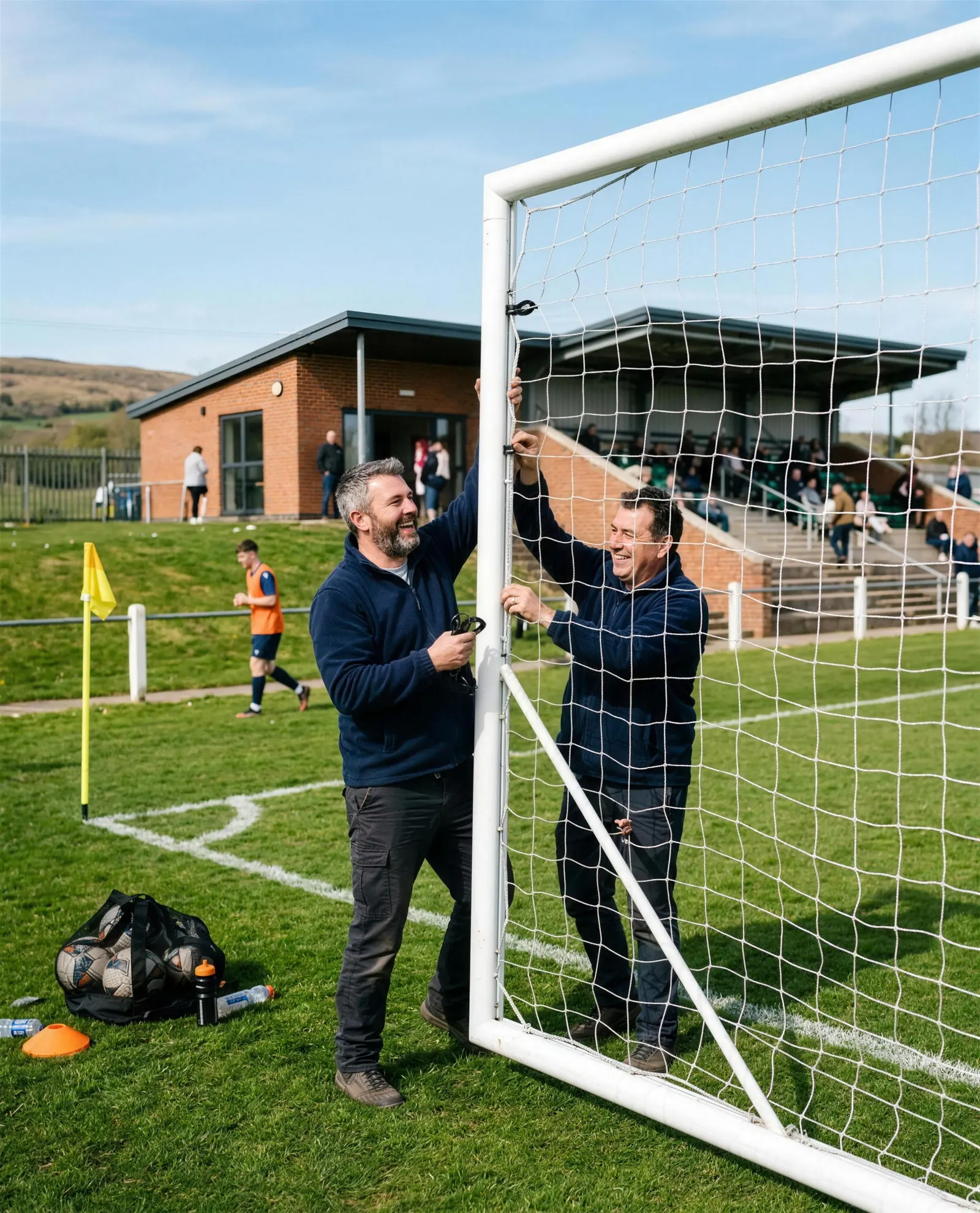 Two club volunteers in navy fleeces putting up a full-size goal and clipping on the net, a modern clubhouse and covered stand behind