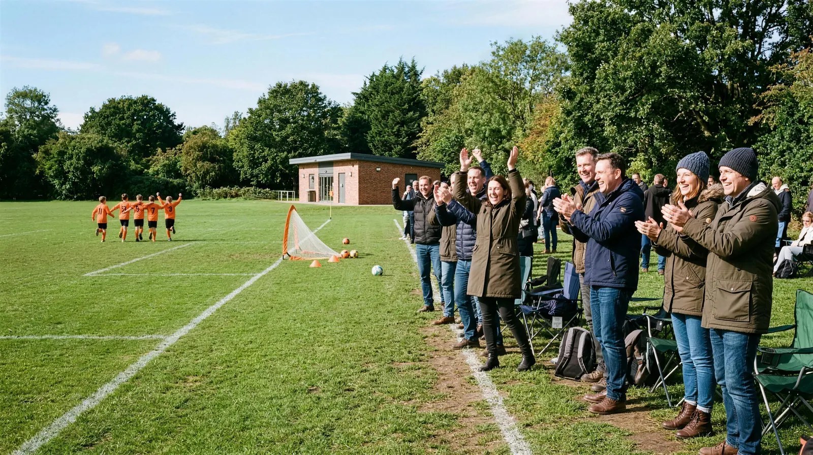 Parents on the touchline of a park pitch cheering and clapping a goal on a sunny morning, young players in orange celebrating in the distance